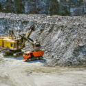 Huge excavator, truck and man standing next on granite quarry. Yellow carver machine, excavator, stonecutter for industrial granite stands in a quarry near granite stones